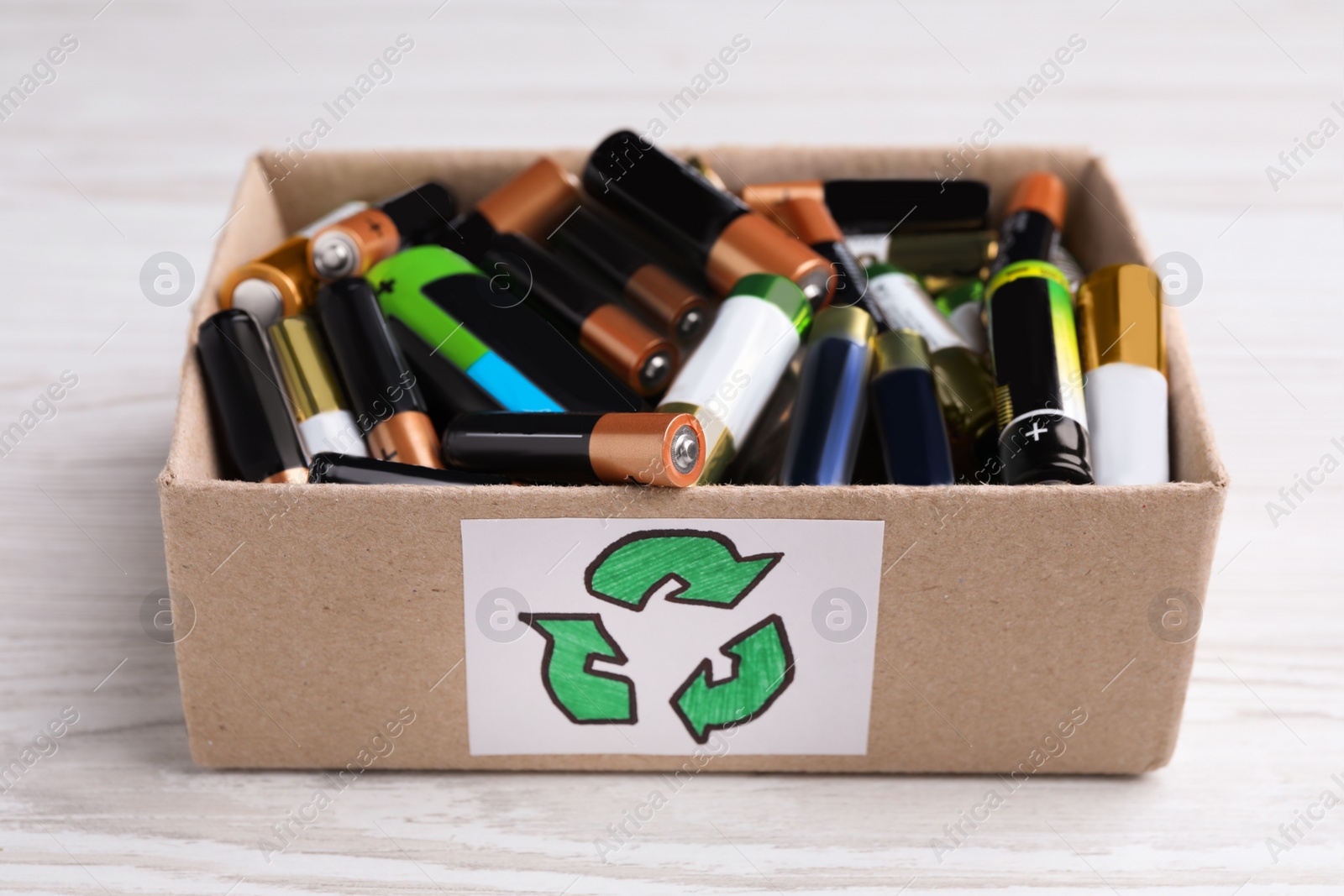 Image of Used batteries in cardboard box with recycling symbol on white wooden table, closeup