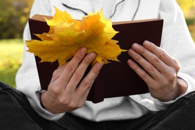 Photo of Woman reading book and holding dry leaves outdoors on autumn day, closeup