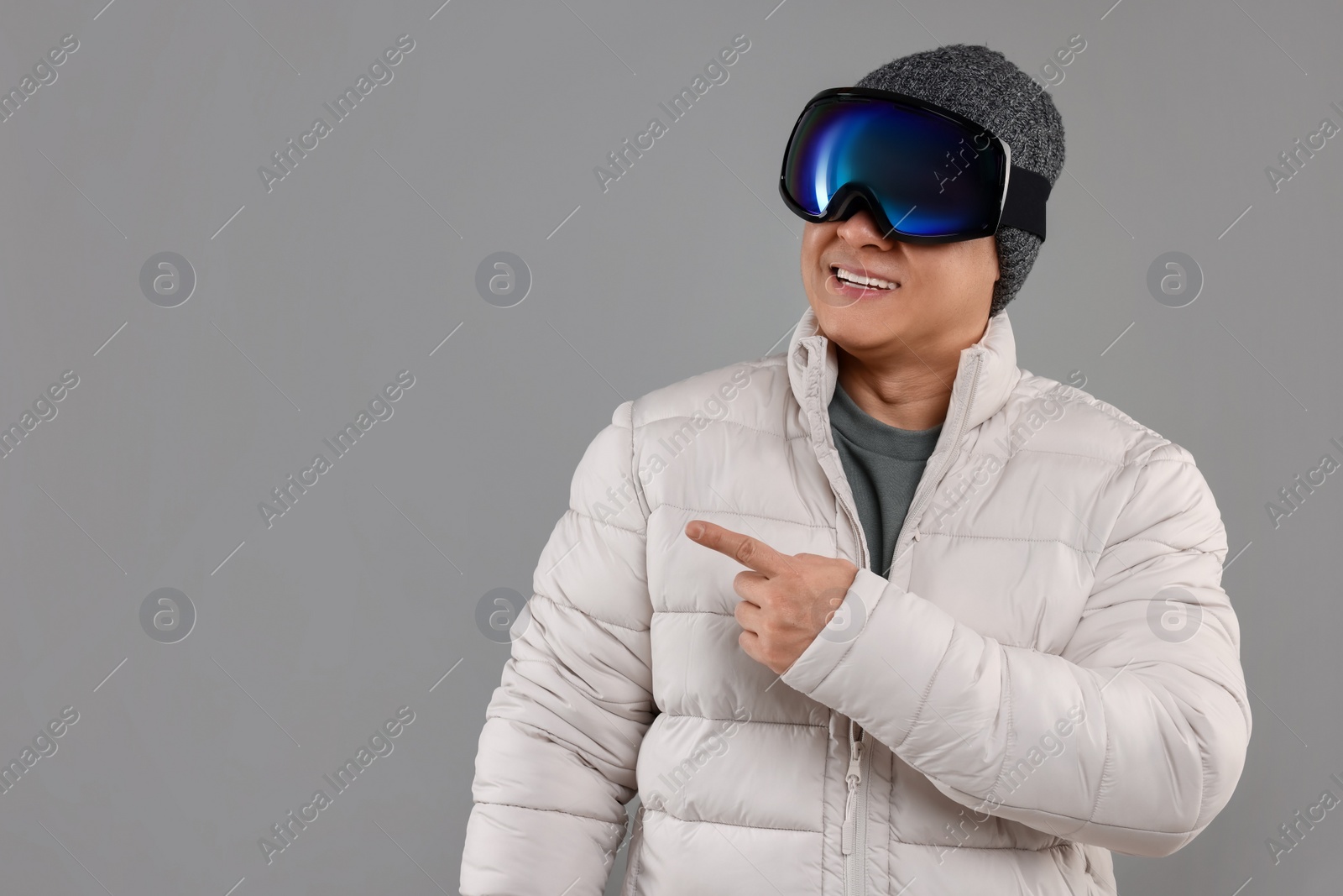 Photo of Winter sports. Happy man in ski goggles pointing at something on grey background. Space for text