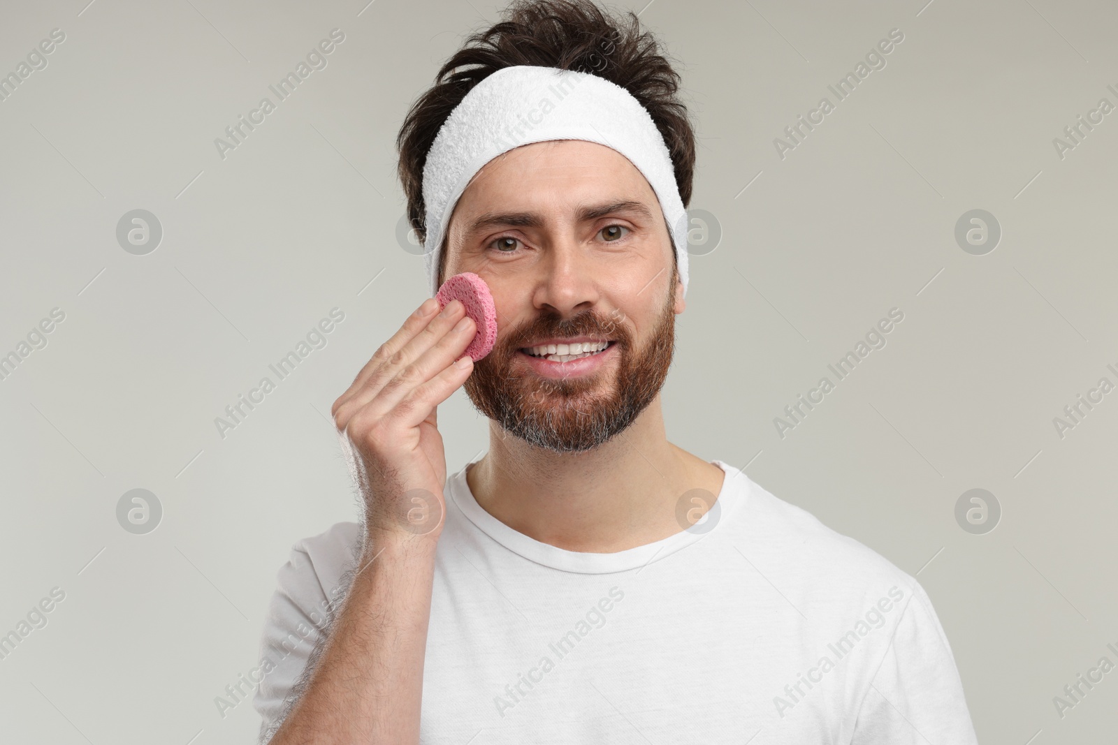 Photo of Man with headband washing his face using sponge on light grey background