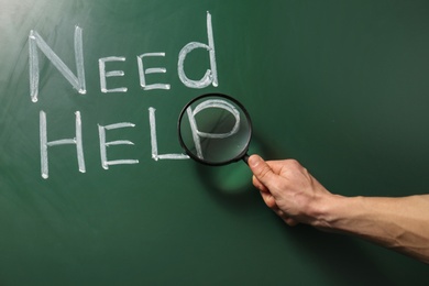 Photo of Man holding magnifying glass over phrase "Need help" written on green chalkboard, closeup