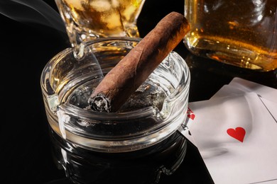 Photo of Smoldering cigar, ashtray, playing cards and whiskey on black mirror surface, closeup