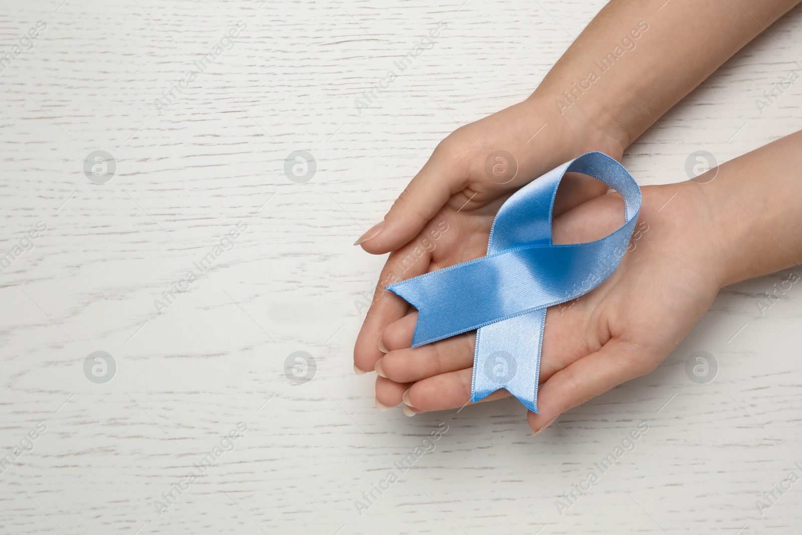 Photo of Woman holding light blue awareness ribbon at white wooden table, top view. Space for text