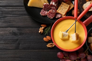 Photo of Pot of tasty cheese fondue and snacks on black wooden table, flat lay. Space for text