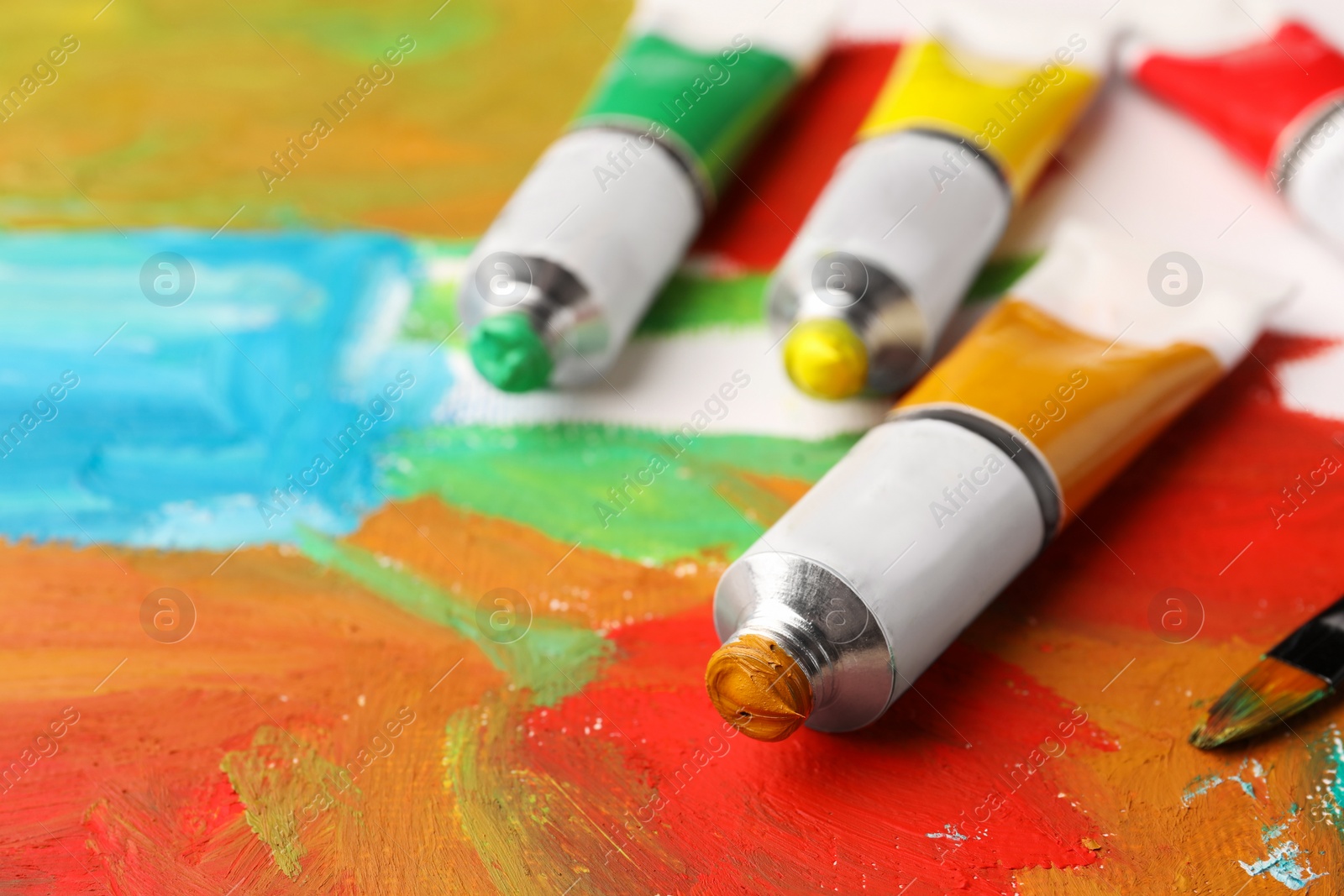 Photo of Tubes of colorful oil paints and brush on canvas with abstract painting, closeup