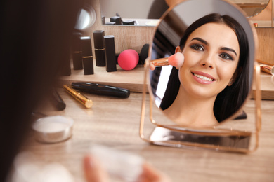 Photo of Beautiful young woman applying makeup near mirror at dressing table