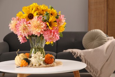 Photo of Beautiful bouquet with bright flowers and small pumpkins on white coffee table near sofa in room, space for text. Autumn atmosphere