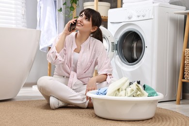 Photo of Happy young housewife with laundry talking on smartphone near washing machine at home