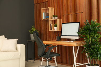 Comfortable workplace with computer near wooden wall in stylish room interior. Home office design