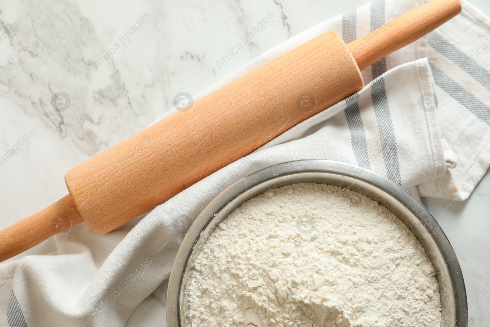 Photo of Flour in bowl, rolling pin and napkin on white marble table, top view