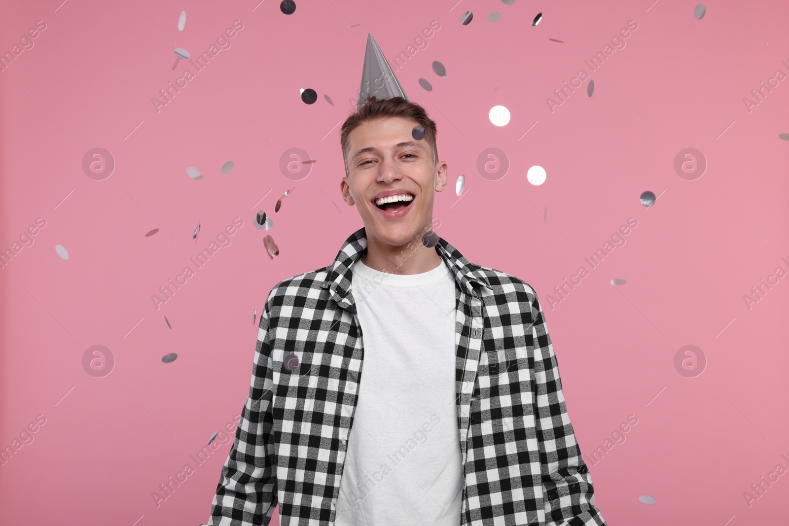 Photo of Happy man in party hat under falling confetti on pink background