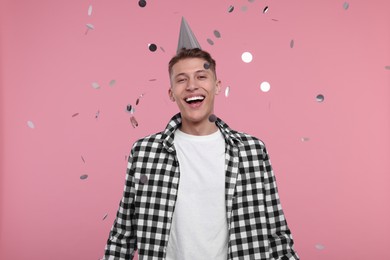 Happy man in party hat under falling confetti on pink background