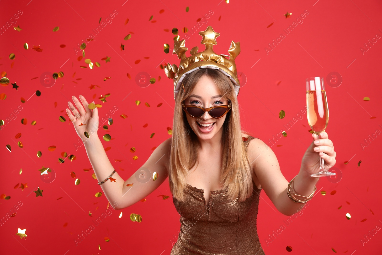 Photo of Happy young woman in party crown with glass of champagne and confetti on red background