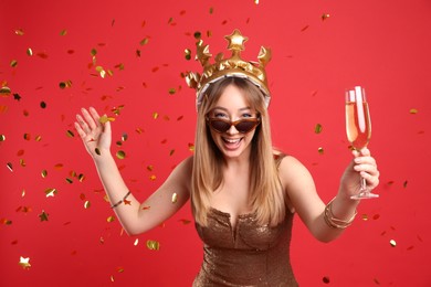 Happy young woman in party crown with glass of champagne and confetti on red background