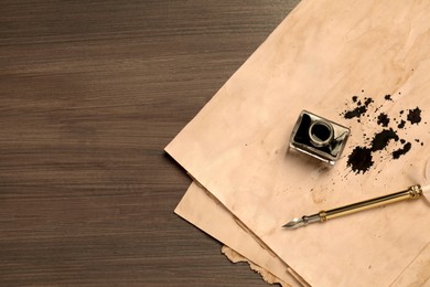 Photo of Fountain pen, inkwell and vintage parchment with ink stains on wooden table, top view. Space for text