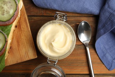 Jar of delicious mayonnaise near fresh sandwich on wooden table, flat lay