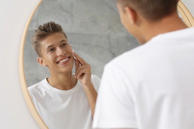 Handsome young man cleaning face with cotton pad near mirror in bathroom