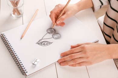 Photo of Woman drawing cherries with graphite pencil in sketchbook at white wooden table, closeup