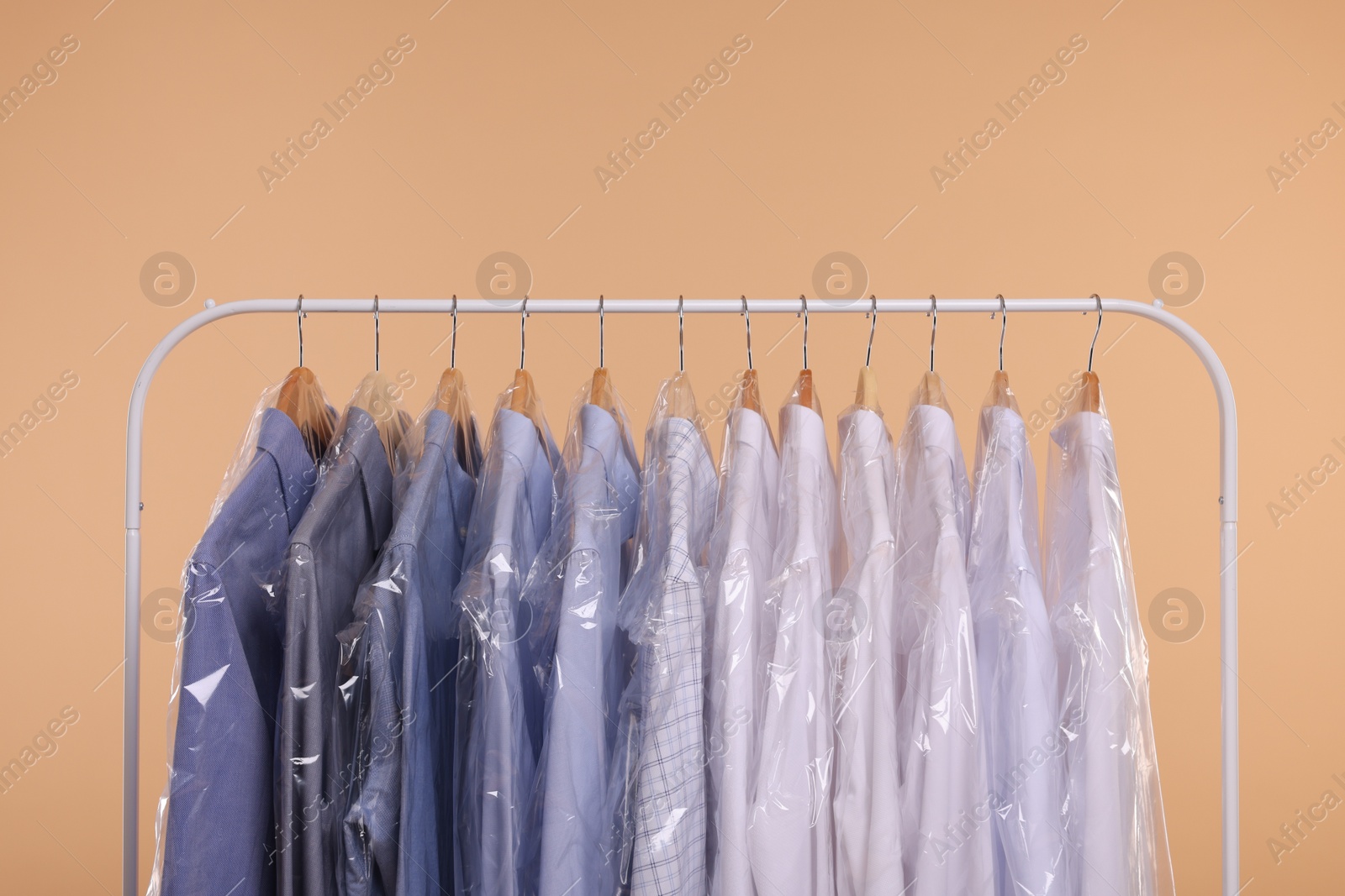 Photo of Dry-cleaning service. Many different clothes in plastic bags hanging on rack against beige background