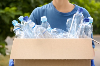 Photo of Woman holding cardboard box with used plastic bottles outdoors, closeup. Recycling problem