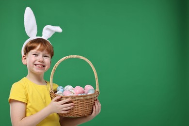 Photo of Easter celebration. Cute little boy with bunny ears and wicker basket full of painted eggs on green background. Space for text