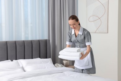 Young maid with stack of towels near bed in hotel room. Space for text