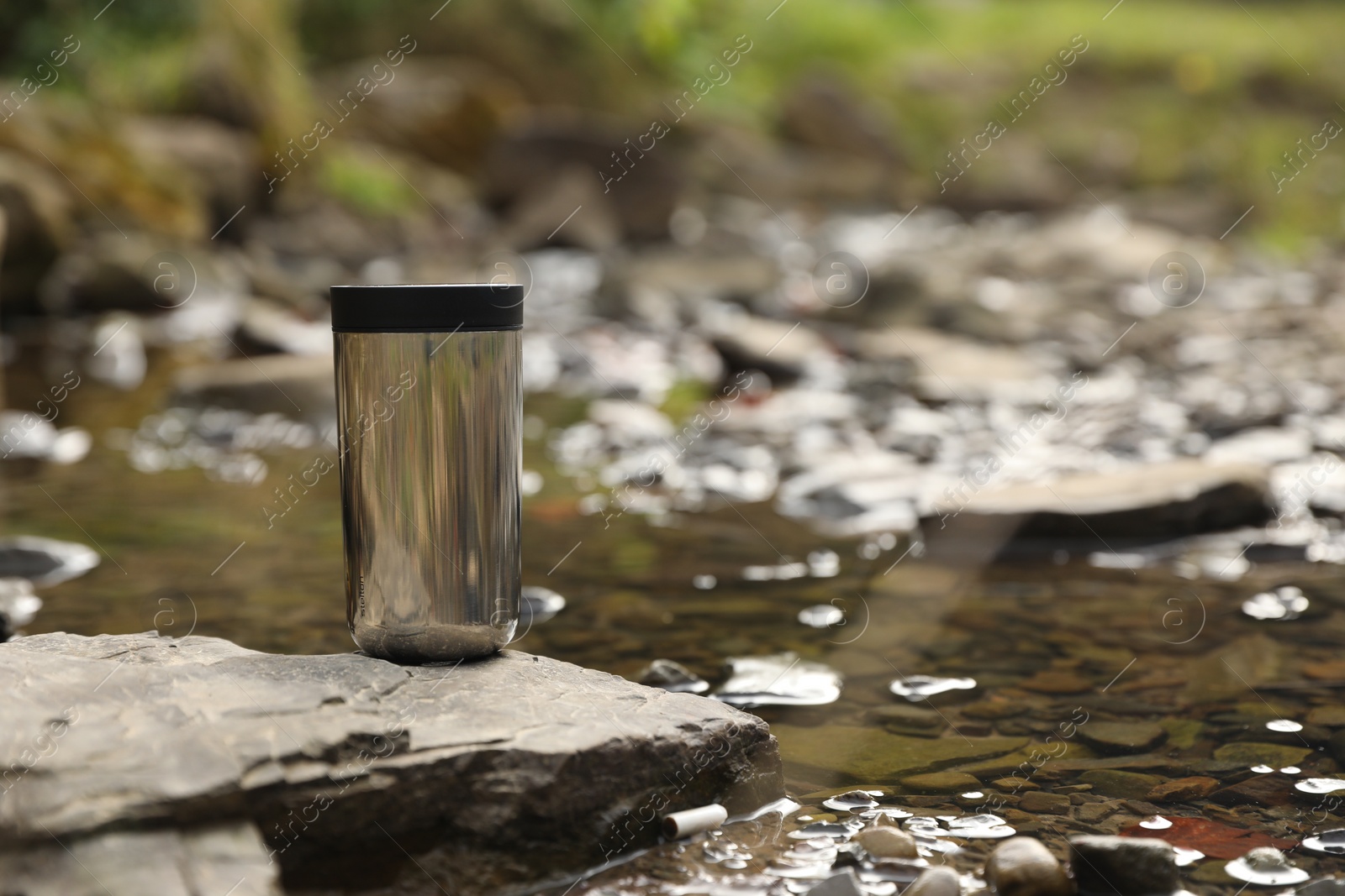 Photo of Metallic thermo tumbler on stone near river, space for text