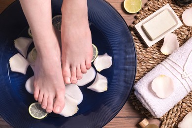 Woman soaking her feet in bowl with water, petals and lime slices on wooden floor, top view. Spa treatment