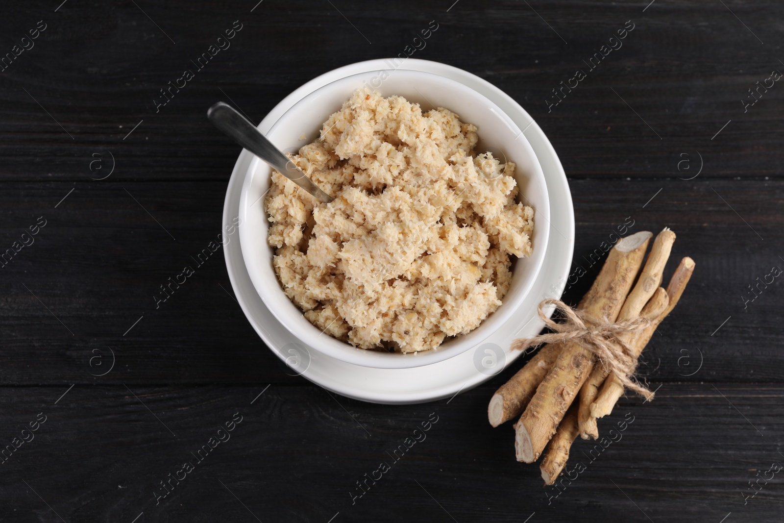 Photo of Bowl of tasty prepared horseradish, spoon and roots on black wooden table, flat lay