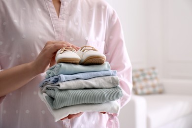 Photo of Woman holding stack of baby's clothes and small shoes indoors, closeup