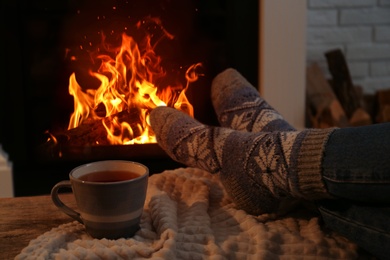 Photo of Woman in warm socks with cup of hot drink resting near fireplace at home, closeup