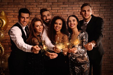 Photo of Happy friends with glasses of sparkling wine and sparklers celebrating New Year indoors