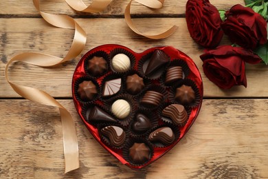 Heart shaped box with delicious chocolate candies, roses and ribbon on wooden table, flat lay