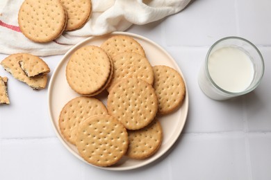 Tasty sandwich cookies and glass of milk on white tiled table, flat lay