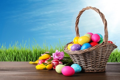 Image of Colorful Easter eggs in wicker basket and spring tulips on wooden table outdoors, space for text 