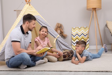Father reading book to children near toy wigwam at home
