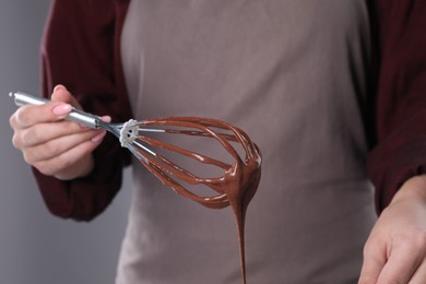 Photo of Woman holding whisk with flowing chocolate cream on grey background, closeup
