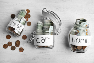 Glass jars with money and tags REST, CAR, HOME on white table, flat lay