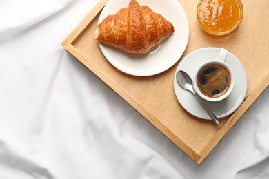 Top view of wooden tray with coffee, croissant and jam on white fabric, space for text. Breakfast in bed