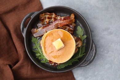 Tasty pancakes with butter, fried bacon and fresh arugula on grey table, top view