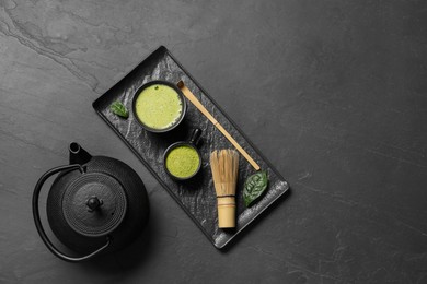 Photo of Cup of fresh matcha tea, bamboo whisk, teapot and green powder on black textured table, flat lay. Space for text