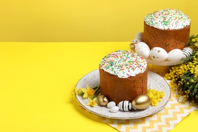 Traditional Easter cakes with sprinkles, painted eggs and beautiful spring flowers on yellow background, space for text