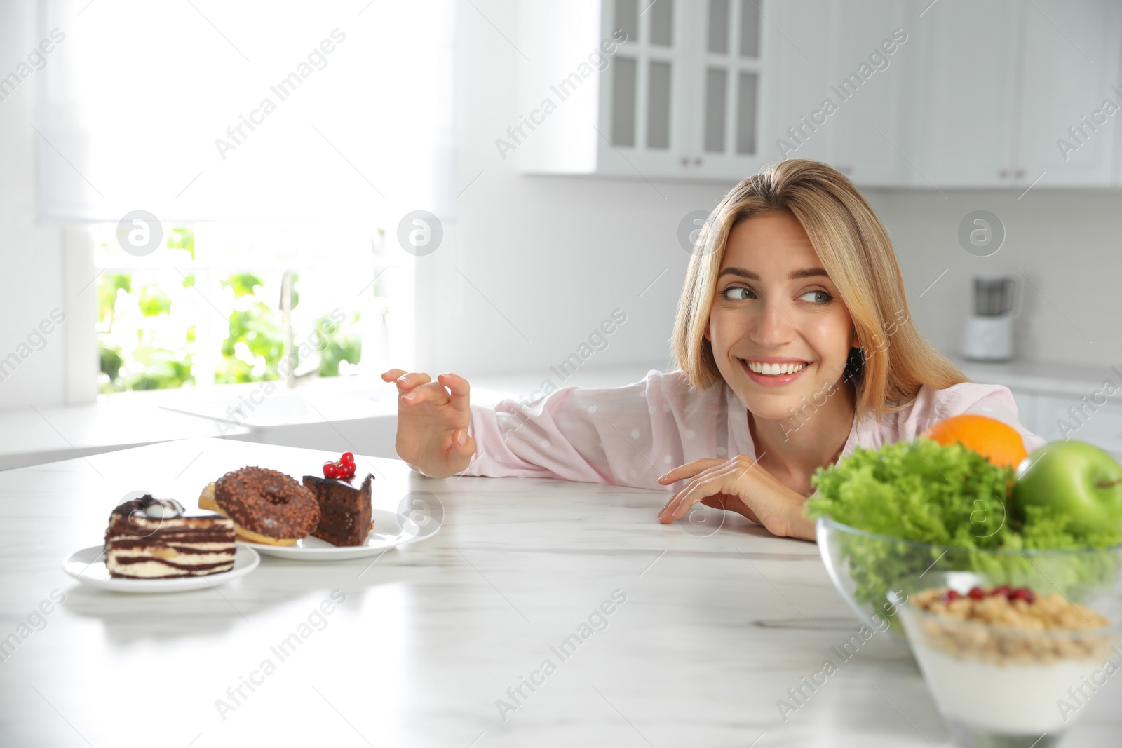 Photo of Woman choosing between sweets and healthy food at white table in kitchen