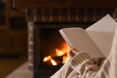 Photo of Woman reading book near burning fireplace at home, closeup