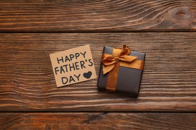 Photo of Card with phrase Happy Father's Day and gift box on wooden background, flat lay
