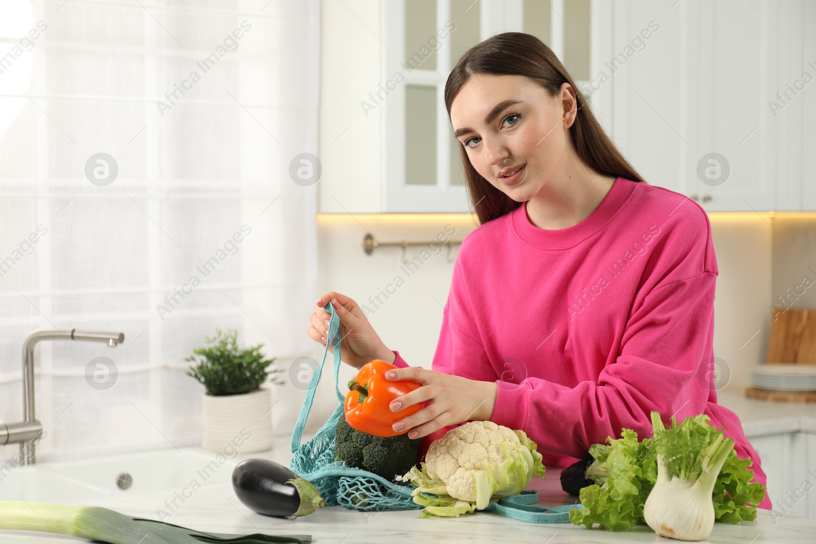 Photo of Woman taking pepper out from string bag at light marble table in kitchen