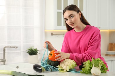 Photo of Woman taking pepper out from string bag at light marble table in kitchen