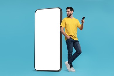 Image of Man with mobile phone standing near huge device with empty screen on light blue background. Mockup for design