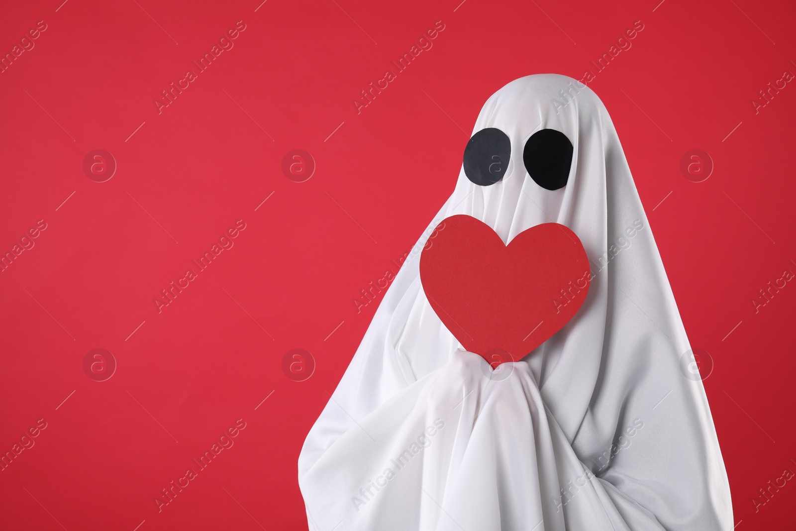 Photo of Cute ghost. Person covered with white sheet holding heart on red background, space for text