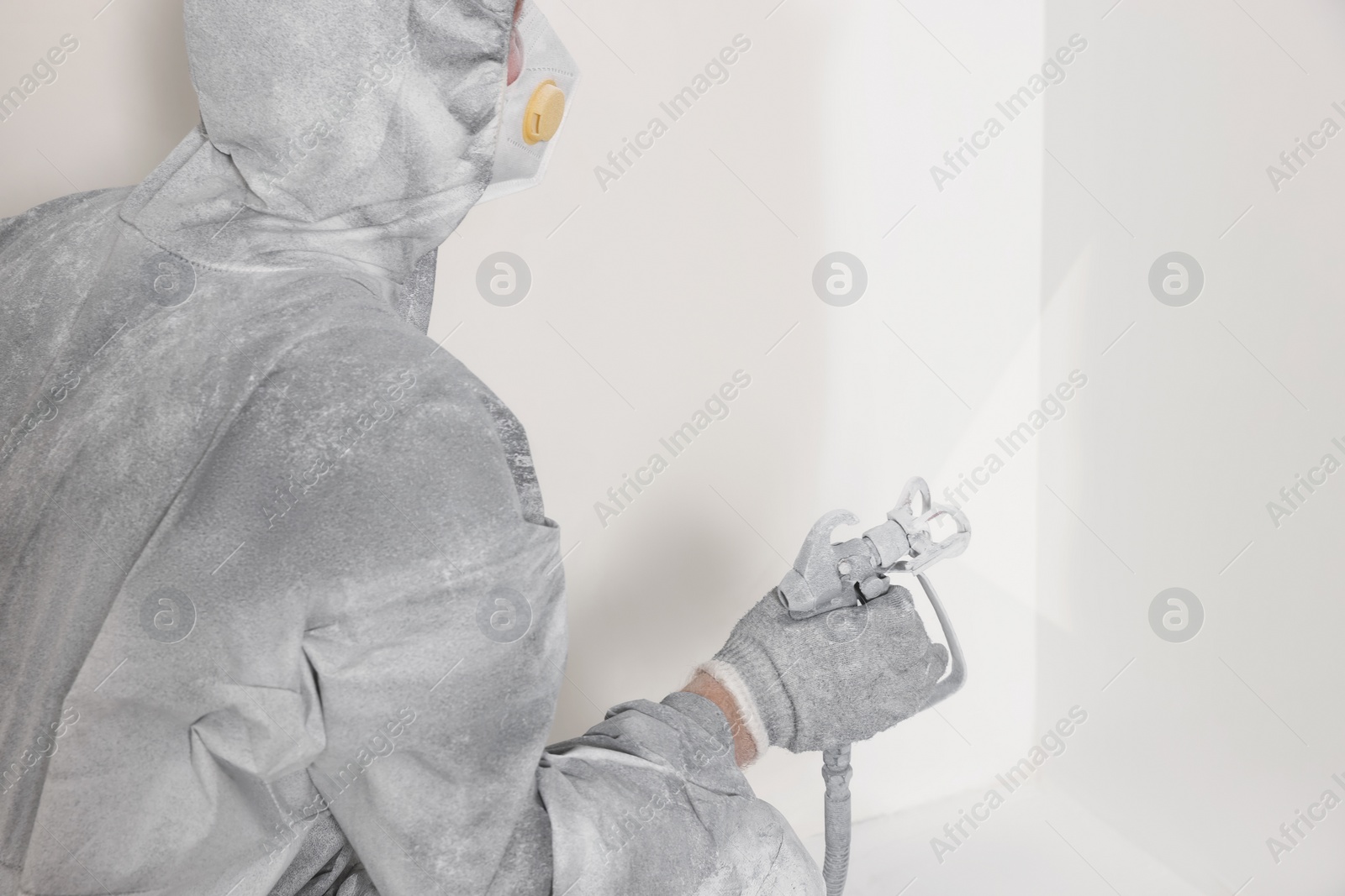 Photo of Decorator in protective overalls painting wall with spray gun indoors, closeup